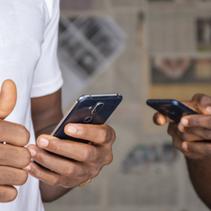 Smartphone Buying Guide 3rd blog image Smartphone Buying Guide Two people holding smartphones, one giving a thumbs-up. They are standing close together, wearing casual shirts. The background shows blurred newspapers.