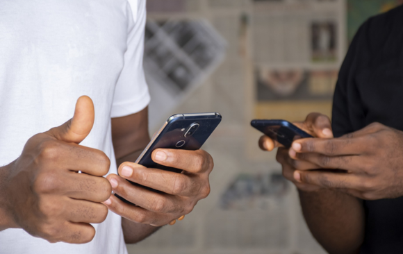 Smartphone Buying Guide 3rd blog image Smartphone Buying Guide Two people holding smartphones, one giving a thumbs-up. They are standing close together, wearing casual shirts. The background shows blurred newspapers.