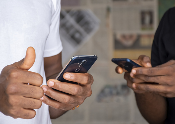 Smartphone Buying Guide Two people holding smartphones, one giving a thumbs-up. They are standing close together, wearing casual shirts. The background shows blurred newspapers.