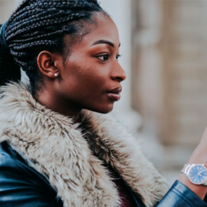 leading smartphone store in Lusaka 1st blog Image A woman with braided hair and a faux fur-collared jacket holds a smartphone, focused and serious. She stands outdoors, conveying concentration and engagement.