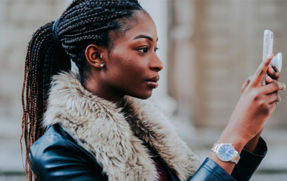 leading smartphone store in Lusaka 1st blog Image A woman with braided hair and a faux fur-collared jacket holds a smartphone, focused and serious. She stands outdoors, conveying concentration and engagement.