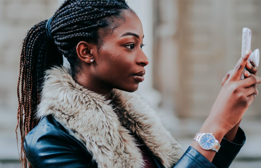 leading smartphone store in Lusaka 1st blog Image A woman with braided hair and a faux fur-collared jacket holds a smartphone, focused and serious. She stands outdoors, conveying concentration and engagement.