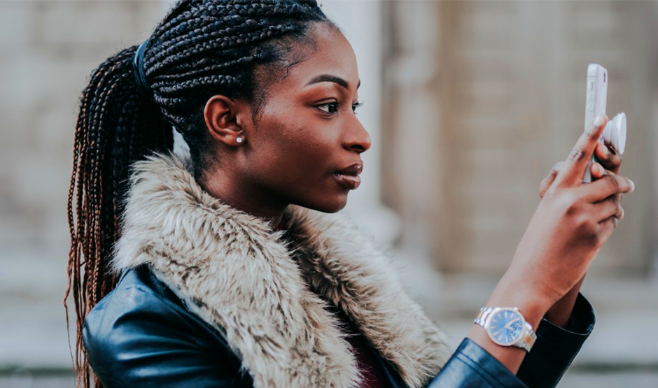 leading smartphone store in Lusaka 1st blog Image A woman with braided hair and a faux fur-collared jacket holds a smartphone, focused and serious. She stands outdoors, conveying concentration and engagement.